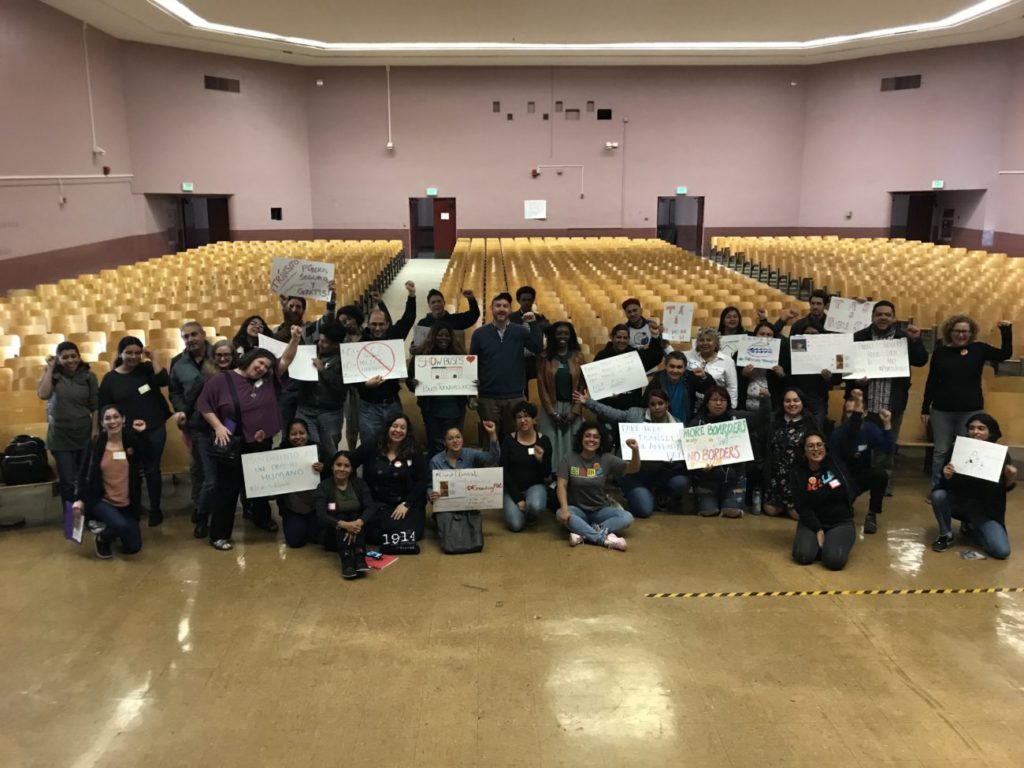 A group of people smiling and raising their fists. They are on the stage facing away from the theater seats and holding up posters.