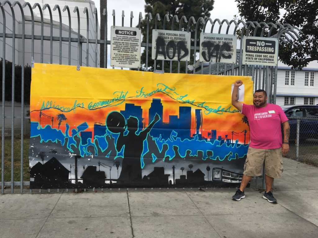 A photo of a man standing in front of a banner with his fist raised. The banner has a silhouette of the Los Angeles skyline painted on it, with silhouettes of people around. In cursive on the top it says The Alliance for Community Transit Los Angeles.