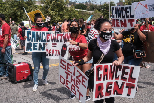 A group of masked activists with signs that say Cancel Rent, Eviction is Death, Seize the Debt, and Cancel Rent.