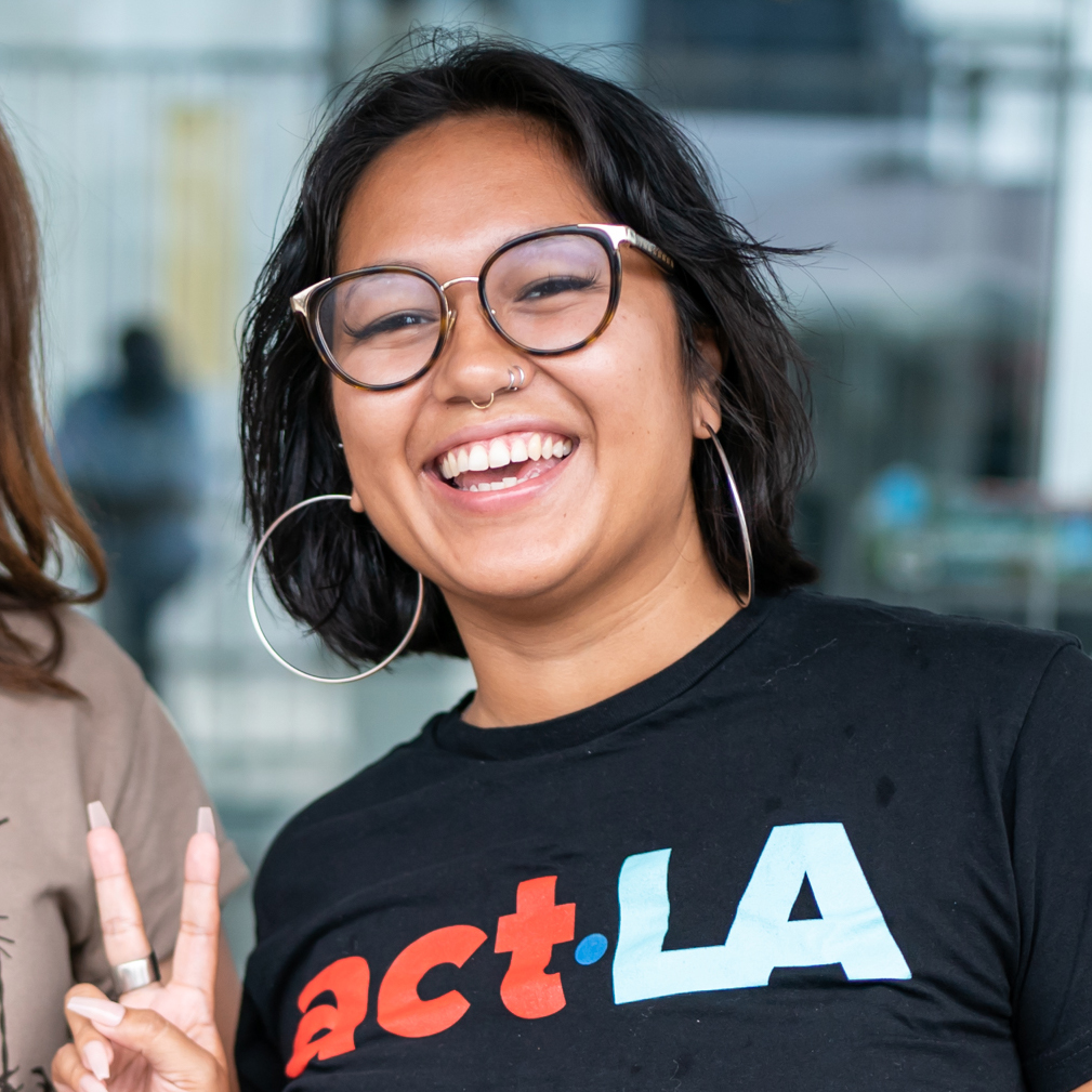 A photo of Carmina Calderon. Her fingers are up in a peace sign and she is smiling while wearing an ACT Los Angeles t-shirt.