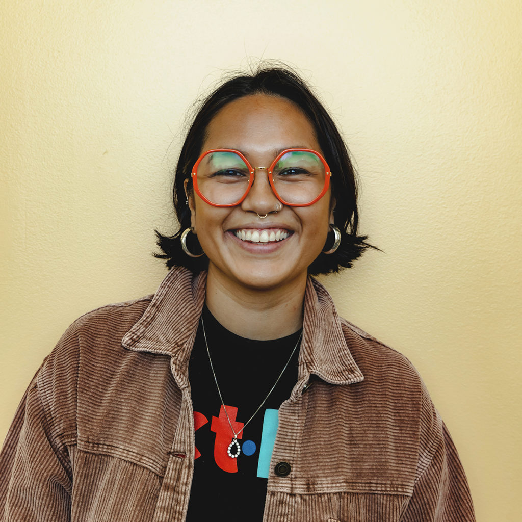 A headshot photo of Carmina Calderon. She has short, dark hair and is smiling with her teeth out. She is wearing large, bright glasses and an ACT Los Angeles t-shirt.
