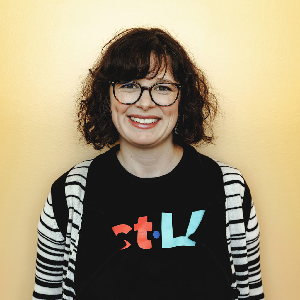 A headshot of Tala Oszkay. She has short, dark, curly hair and is smiling with her teeth. She is wearing dark, round glasses and an ACT Los Angeles t-shirt.