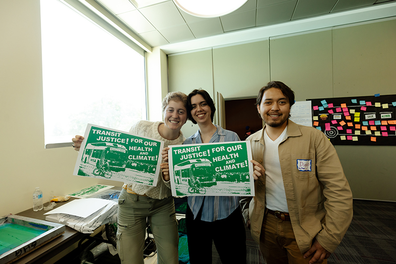 Three smiling people are holding up posters that say "Transit Justice for our health and climate!"