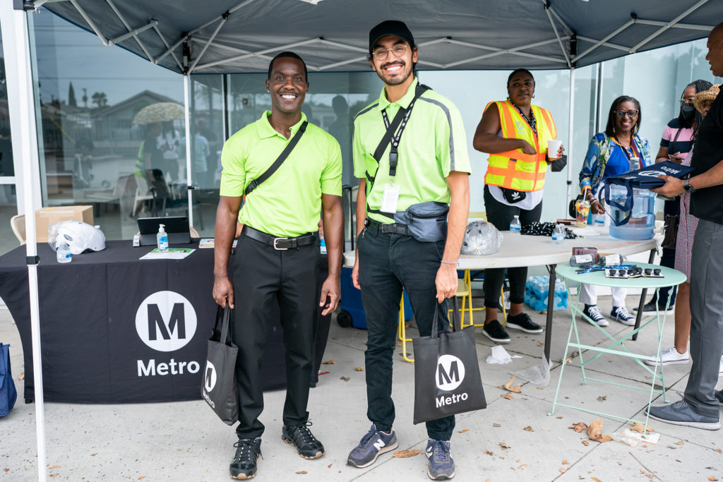 A photo of two Los Angeles Metro transit ambassadors. They are smiling broadly and wearing the bright transit ambassador polos. They are standing in front of a Los Angeles Metro booth with branded merchandise.